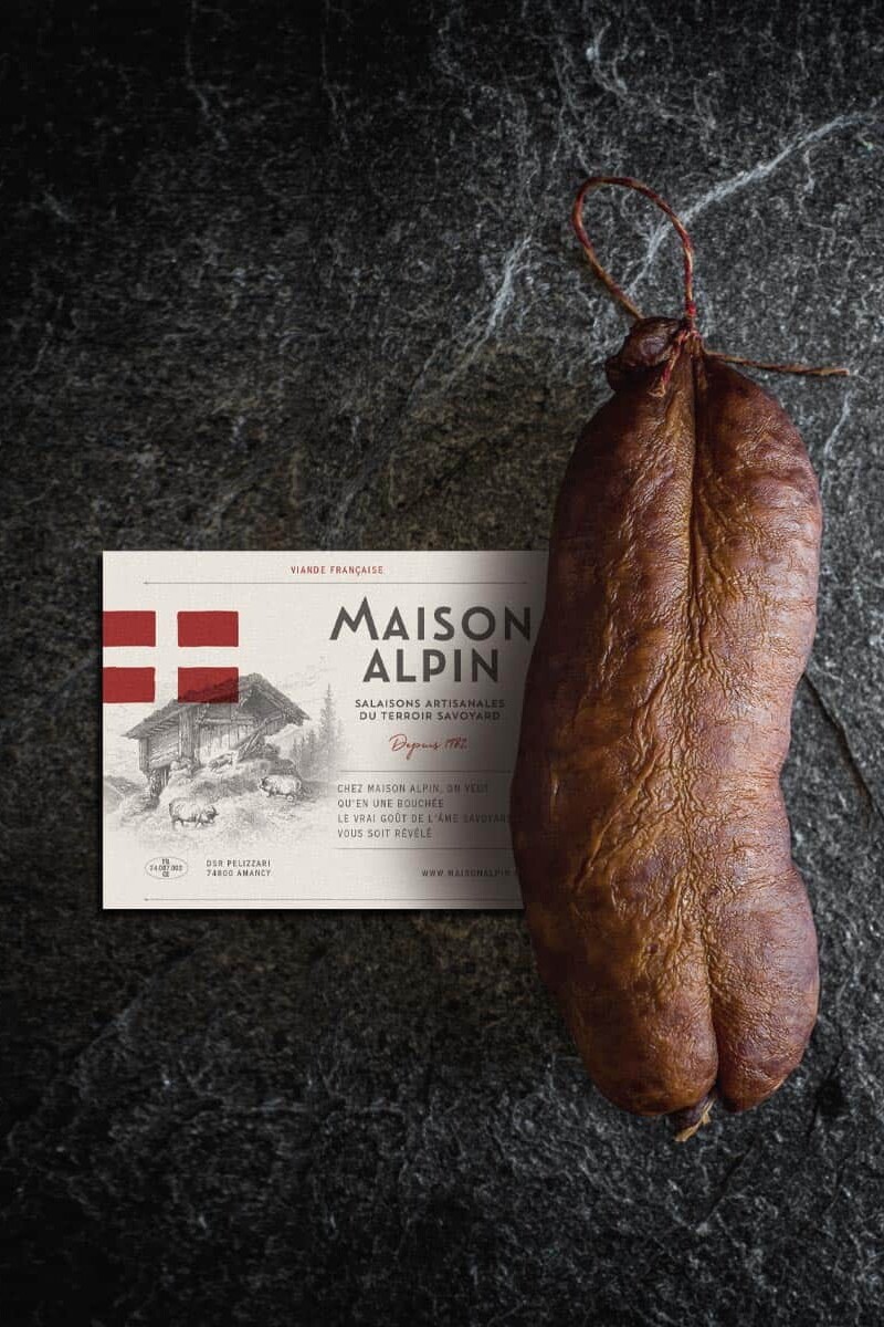 Saucisse s&egrave;che artisanale avec &eacute;tiquette de la Maison Alpin sur fond pierre noire, identit&eacute; visuelle authentique et r&eacute;gionale