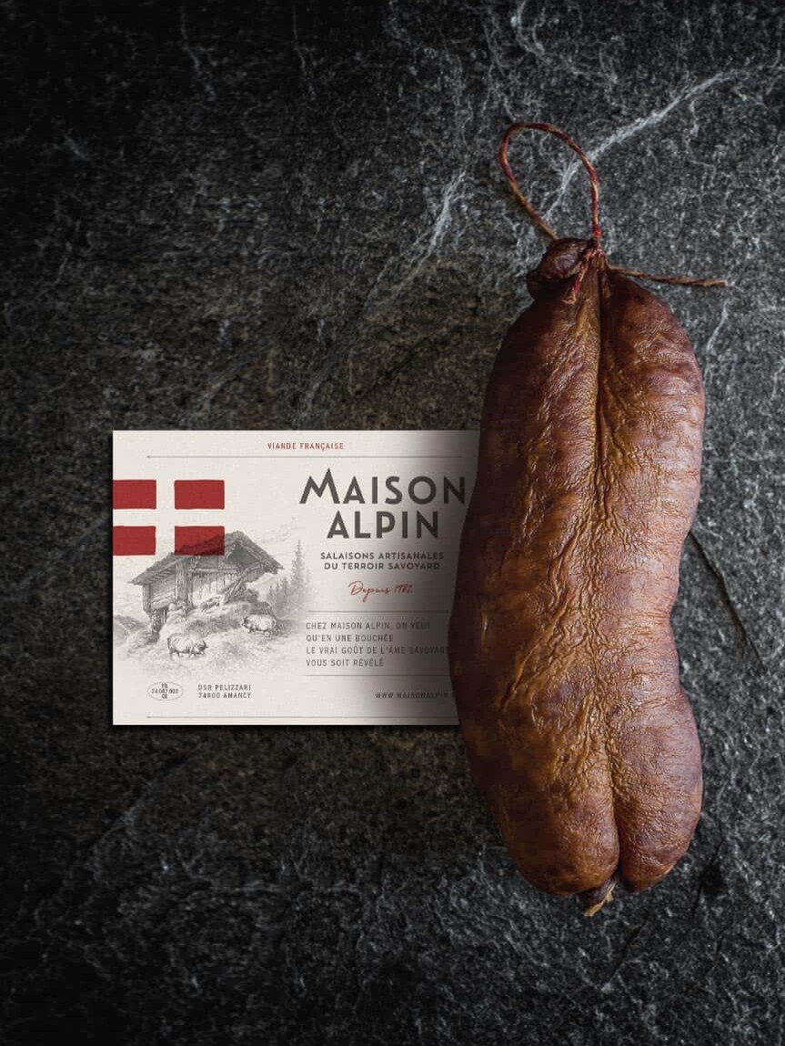 Saucisse s&egrave;che artisanale avec &eacute;tiquette de la Maison Alpin sur fond pierre noire, identit&eacute; visuelle authentique et r&eacute;gionale