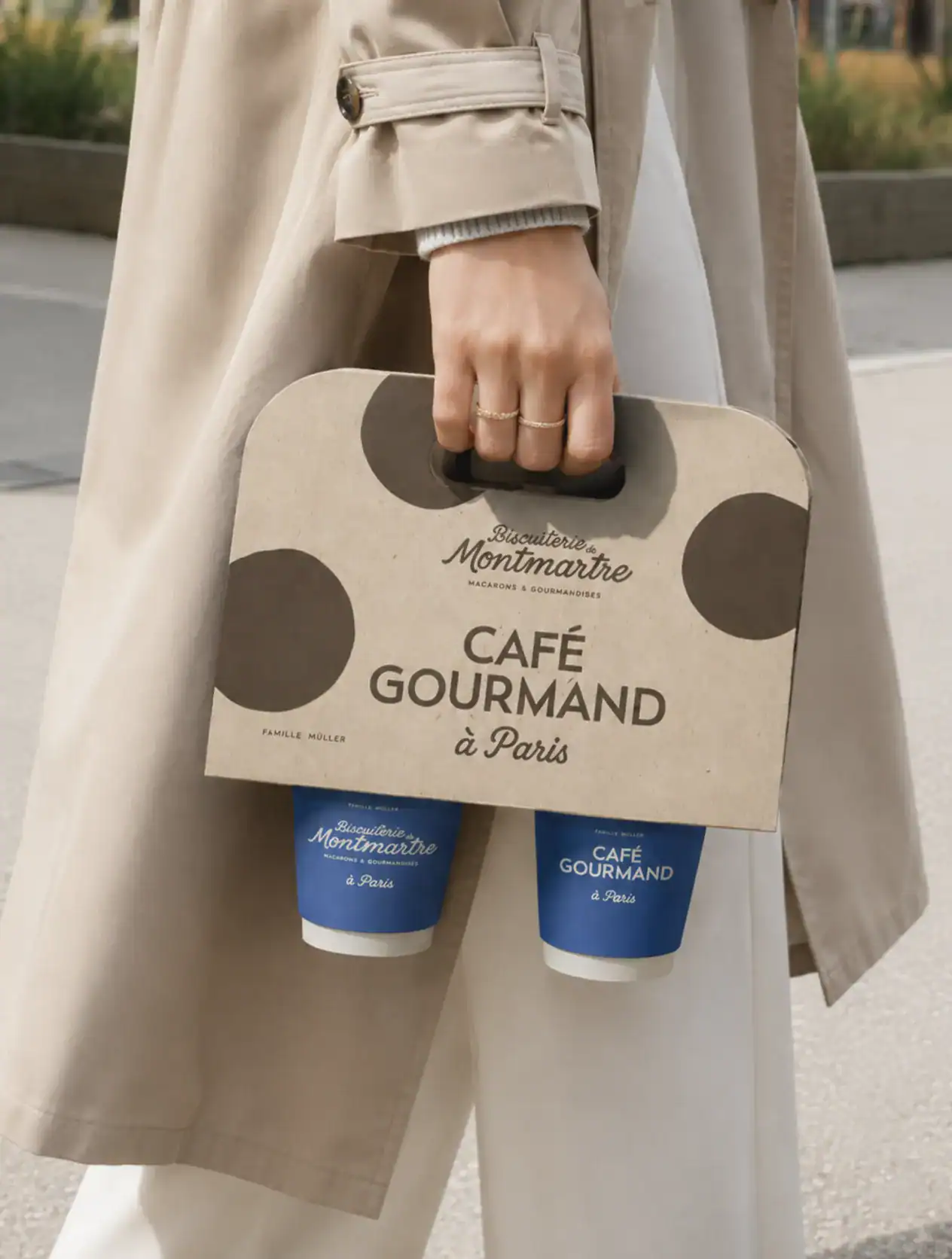 Sac à emporter de la Biscuiterie de Montmartre en carton beige avec logo manuscrit et motif pois-macaron, tenant deux gobelets de café.