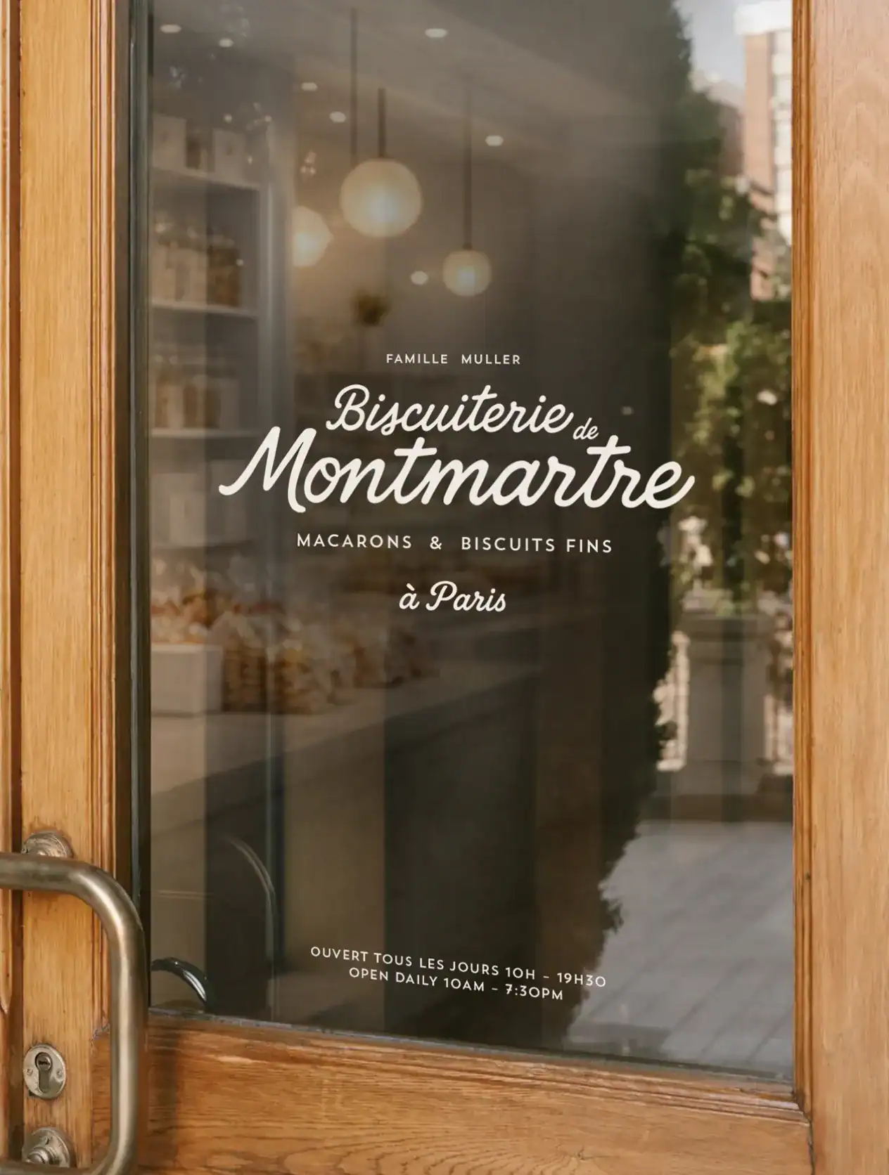 Vitrine de la Biscuiterie de Montmartre avec logo manuscrit, reflet des rues parisiennes et éclairage intérieur chaleureux.