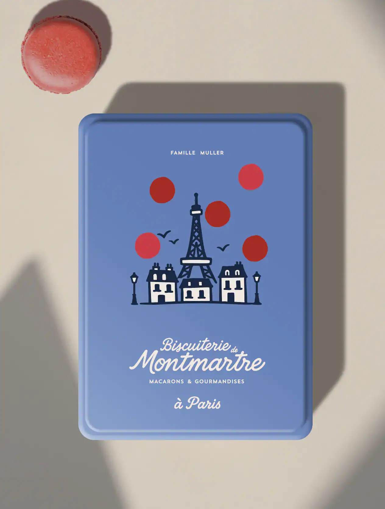 Boîte à macarons de la Biscuiterie de Montmartre, couleur bleu parisien avec motif pois-macaron rouge, logo manuscrit familial et illustration de la tour Eiffel.