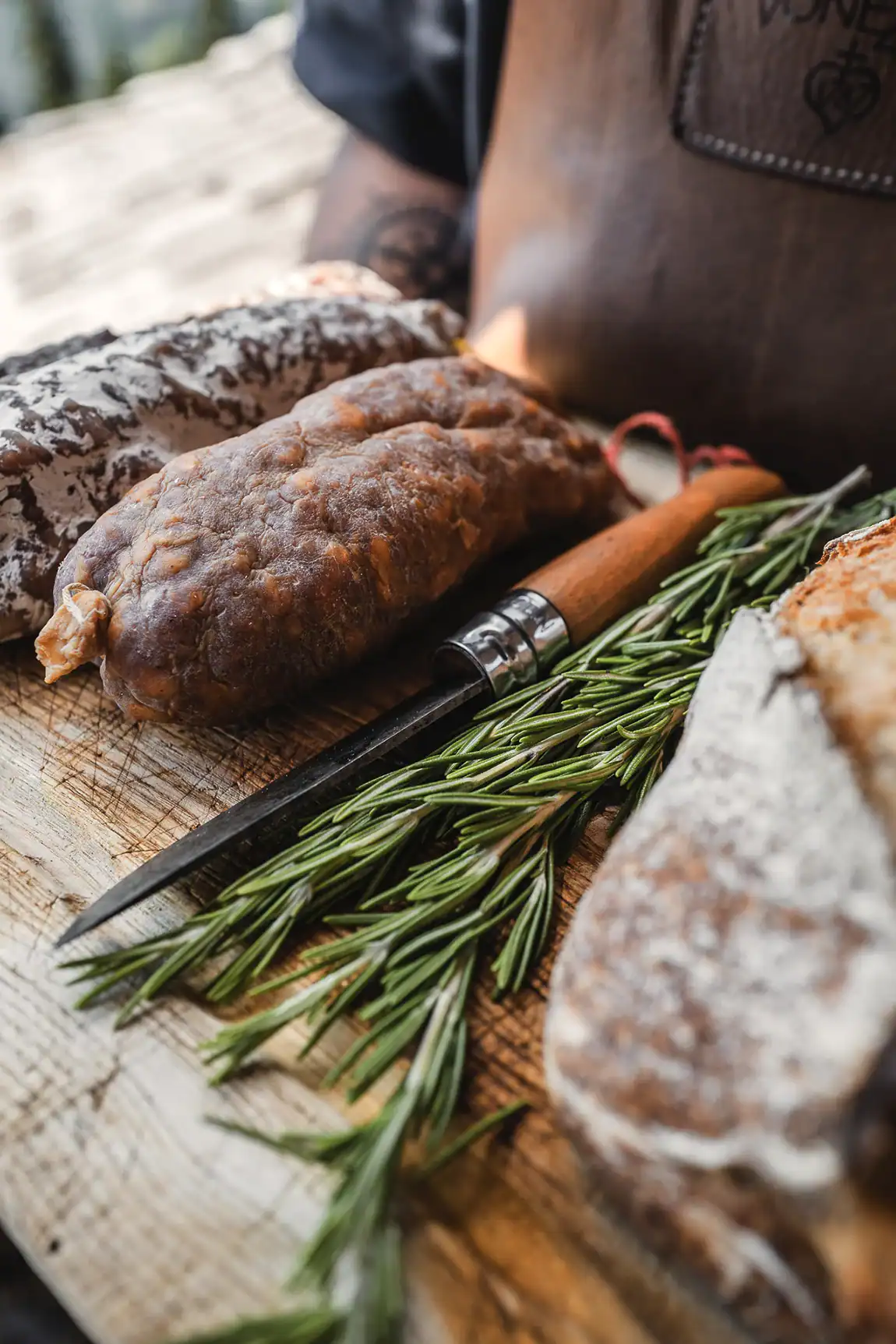 Saucisson sec artisanal Maison Alpin pr&eacute;sent&eacute; avec romarin et couteau sur une planche en bois.