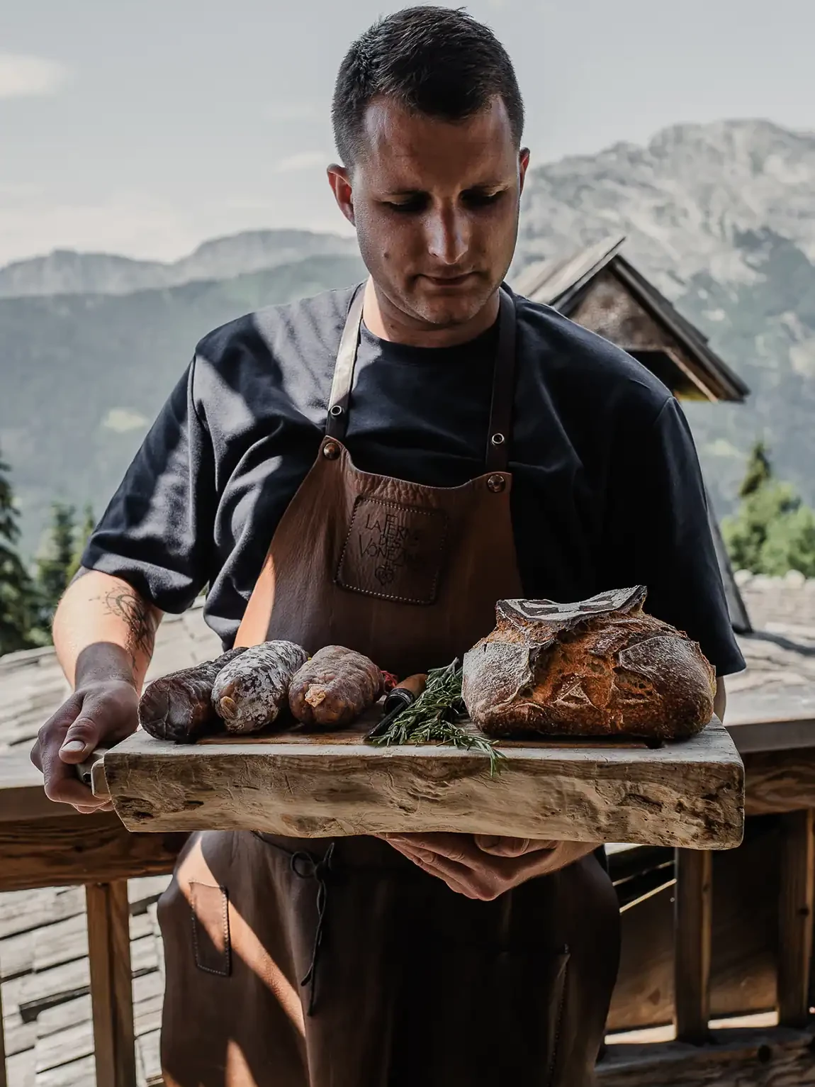 Artisan Maison Alpin portant une planche de saucissons et un pain savoyard devant les montagnes de Haute-Savoie.