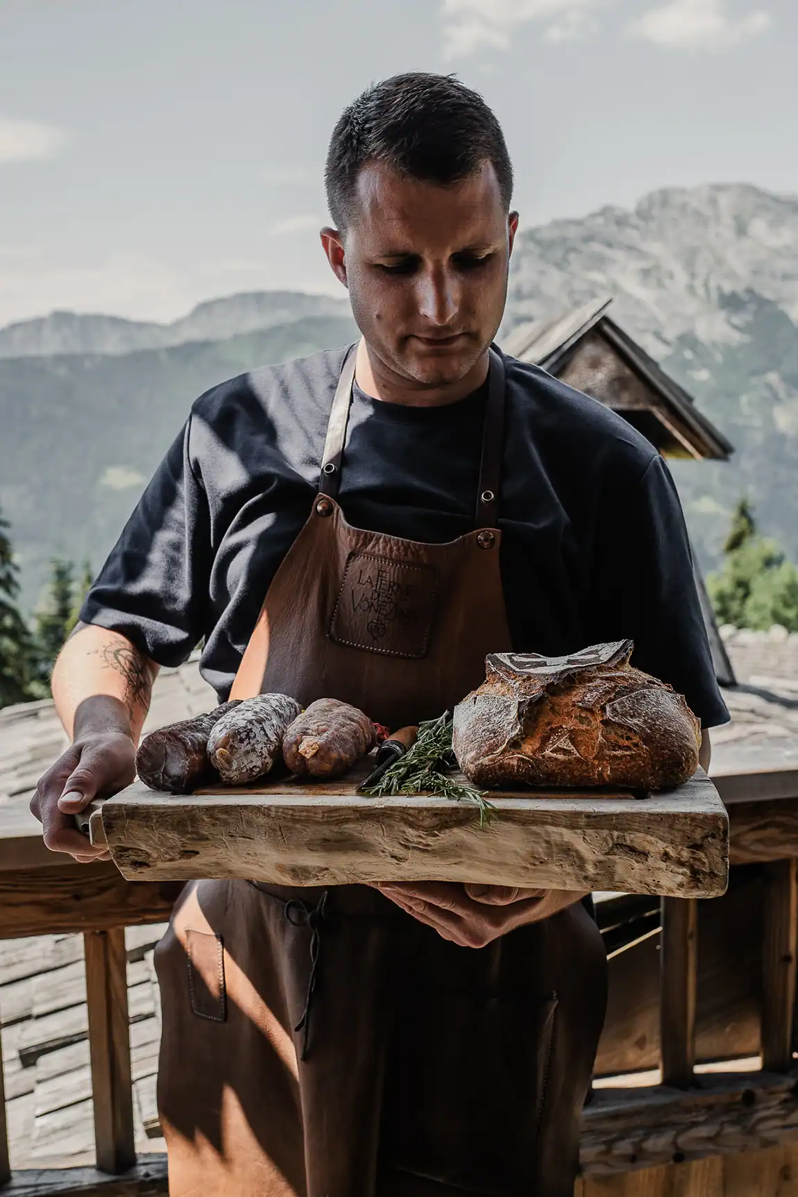 Artisan Maison Alpin portant une planche de saucissons et un pain savoyard devant les montagnes de Haute-Savoie.