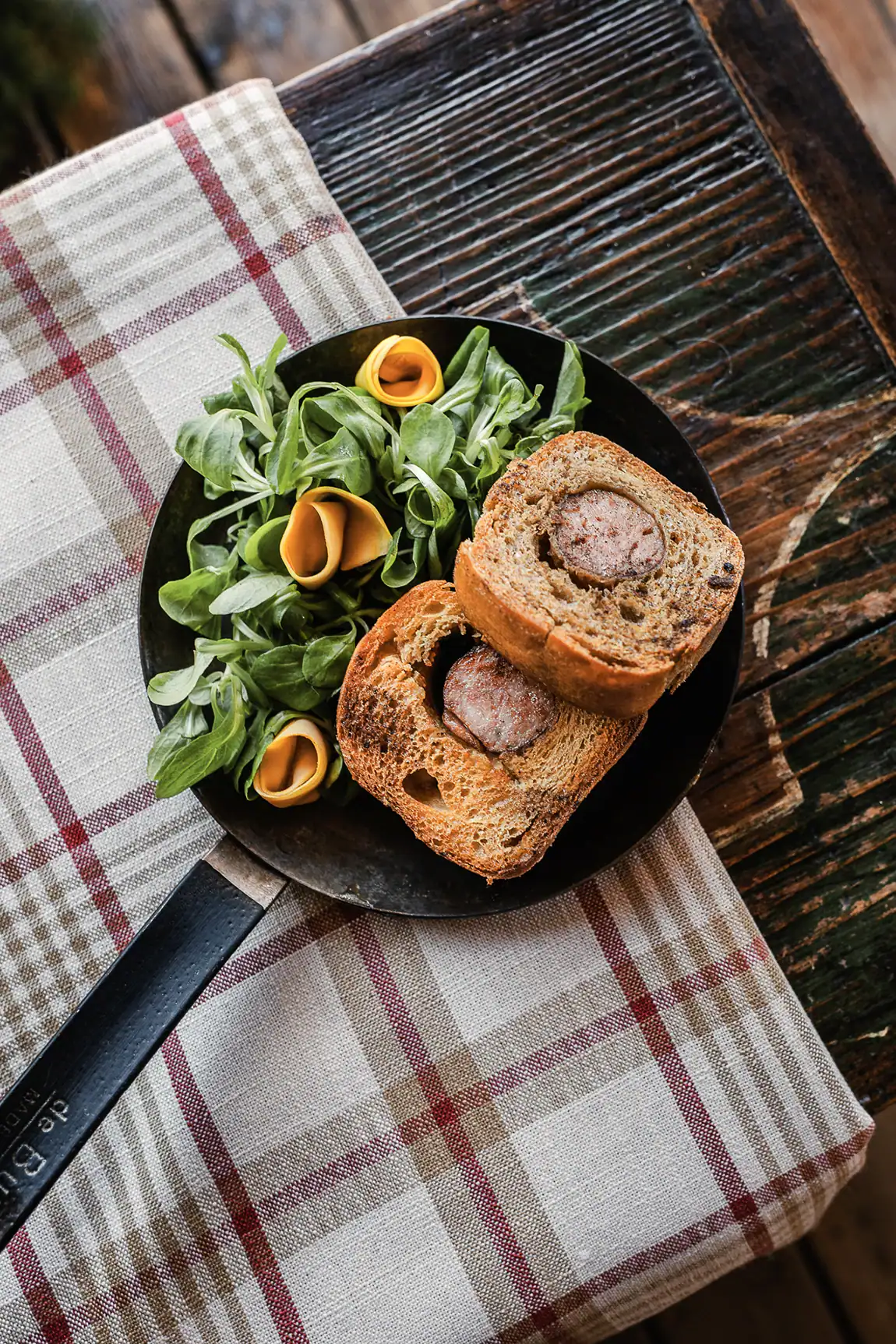 Saucisse artisanale Maison Alpin cuite dans du pain, servie avec salade et l&eacute;gumes marin&eacute;s.